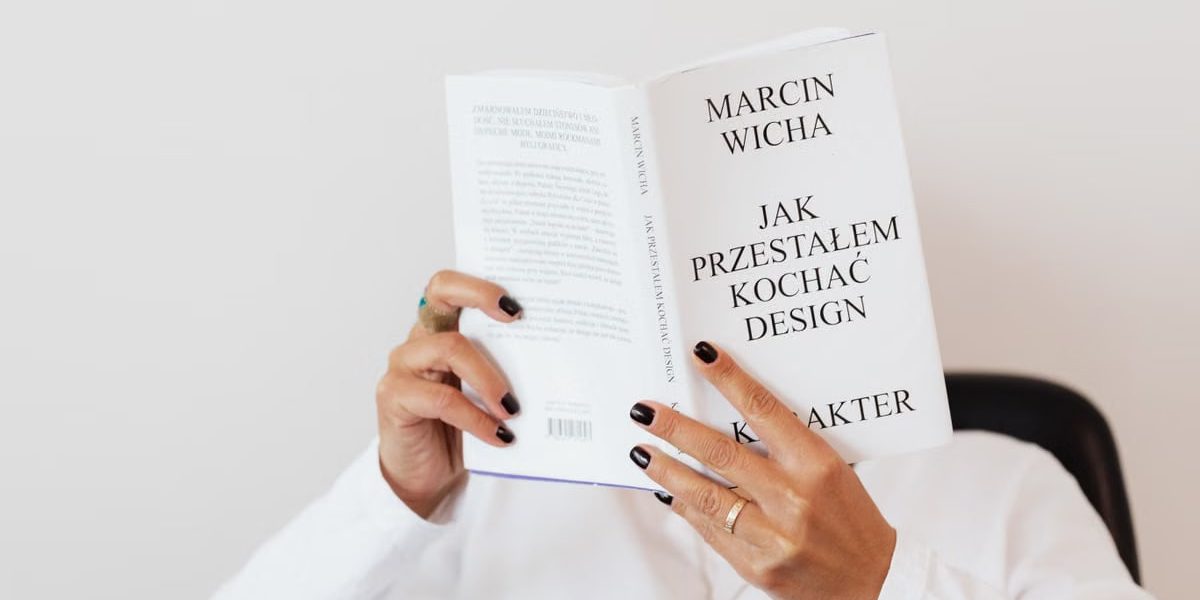 Anonymous female in white shirt with gold chain on neck reading interesting book covering face while sitting on black chair